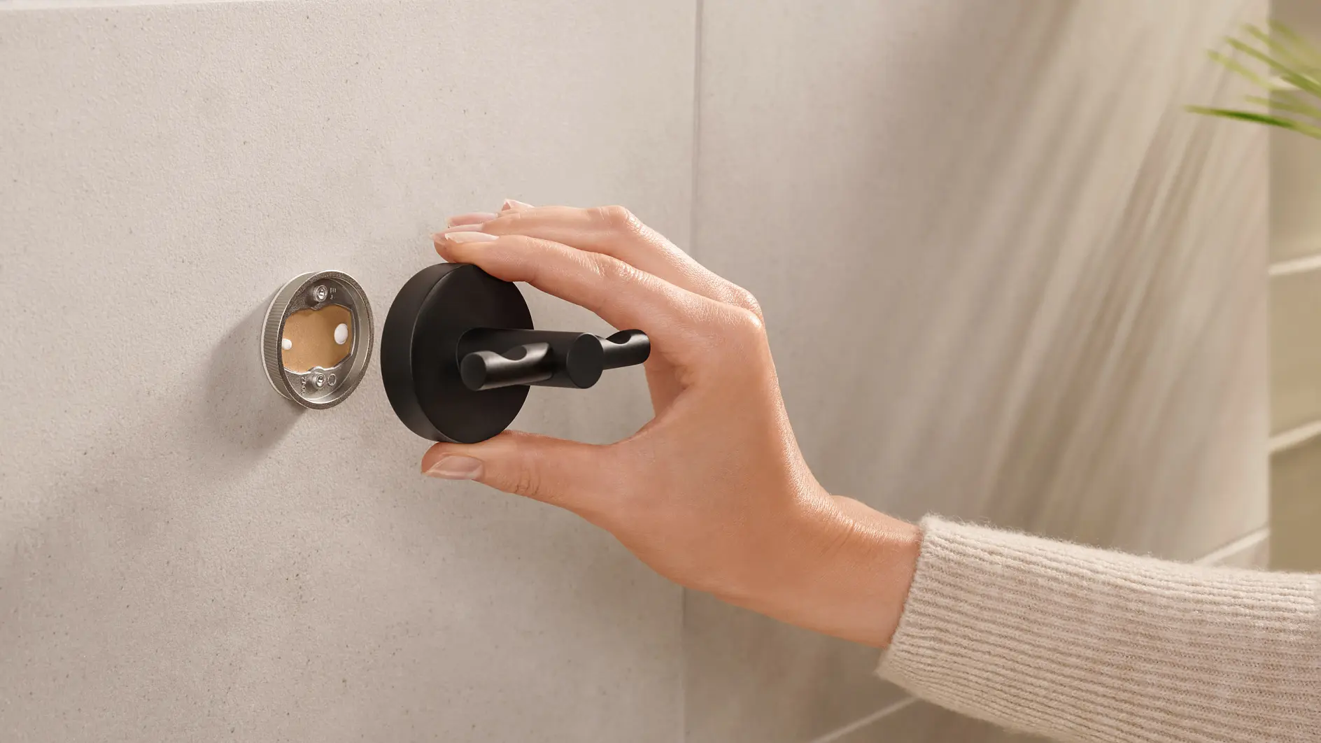 A hand wearing a beige sweater is adjusting a black wall-mounted hook on a light-colored tiled wall using tesa tape. The hand is holding the hook as it aligns with an open circular fitting on the wall. (global.generated_by_ai)