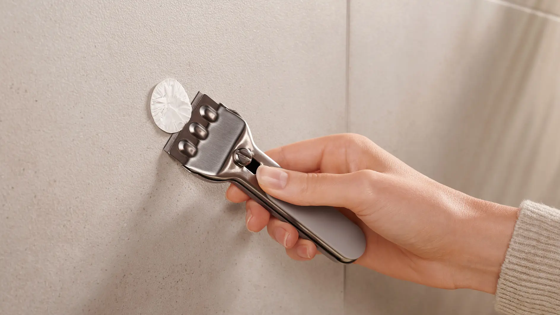 A persons hand holding a metal tool with a grey handle, pressing it against a circular metallic object on a smooth, light-colored vertical surface using tesa tape. (global.generated_by_ai)