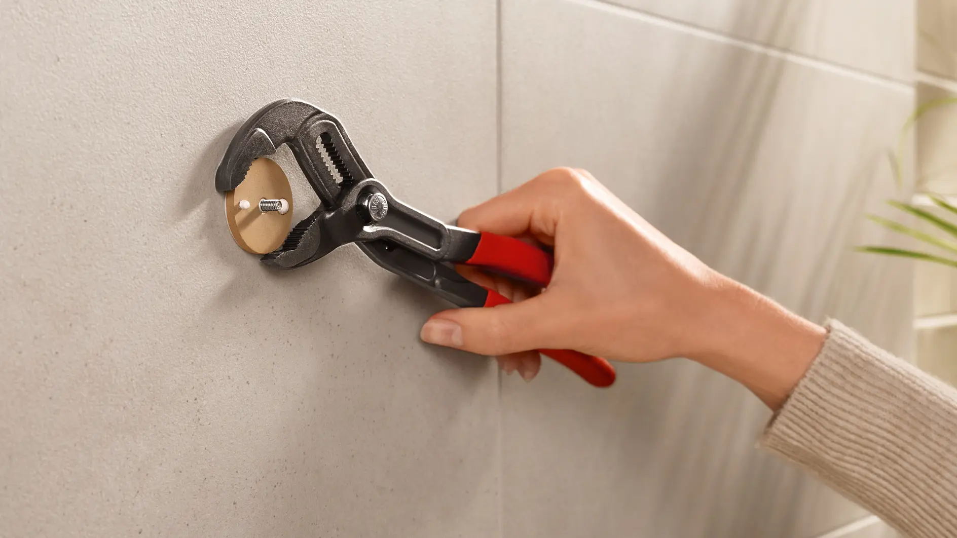 A person is using an adjustable wrench with red handles to tighten or loosen a round fitting on a wall. The wall appears to be tiled in a light gray color, and there are some blurred green leaves visible on the side. (global.generated_by_ai)